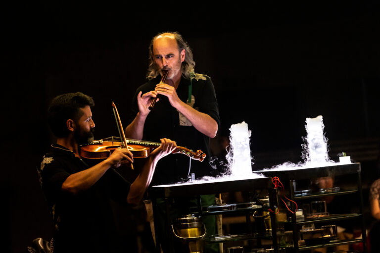 Twee muzikanten optreden op het podium, met een violist en een man die een fluit bespeelt, terwijl waterfonteinen op de achtergrond spuiten.