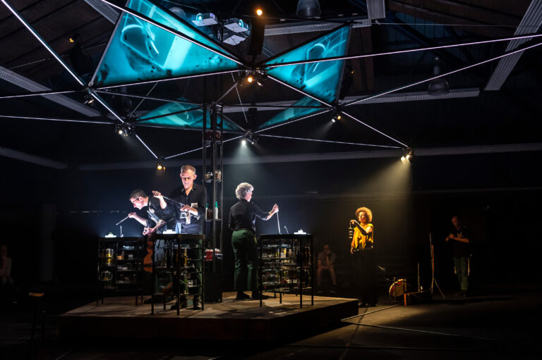 Een silhouet van performers op een podium, verlicht door verschillende lichten en screens met visuele projecties.