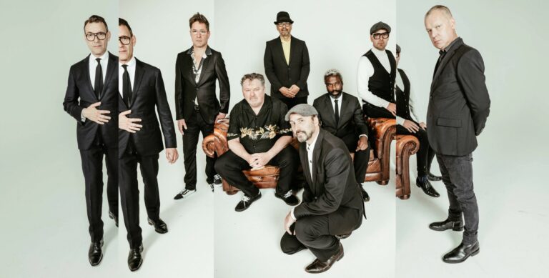 De acht bandleden van New Cool Collective, die allemaal op, voor, naast en achter een bruine bank staan of zitten.
