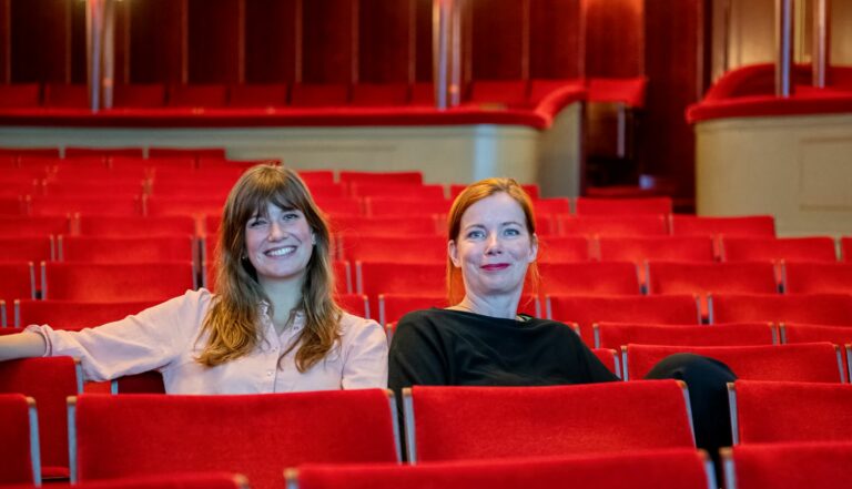 Twee vrouwen zitten in een rijen van rode theaterstoelen met een moderne achtergrond.