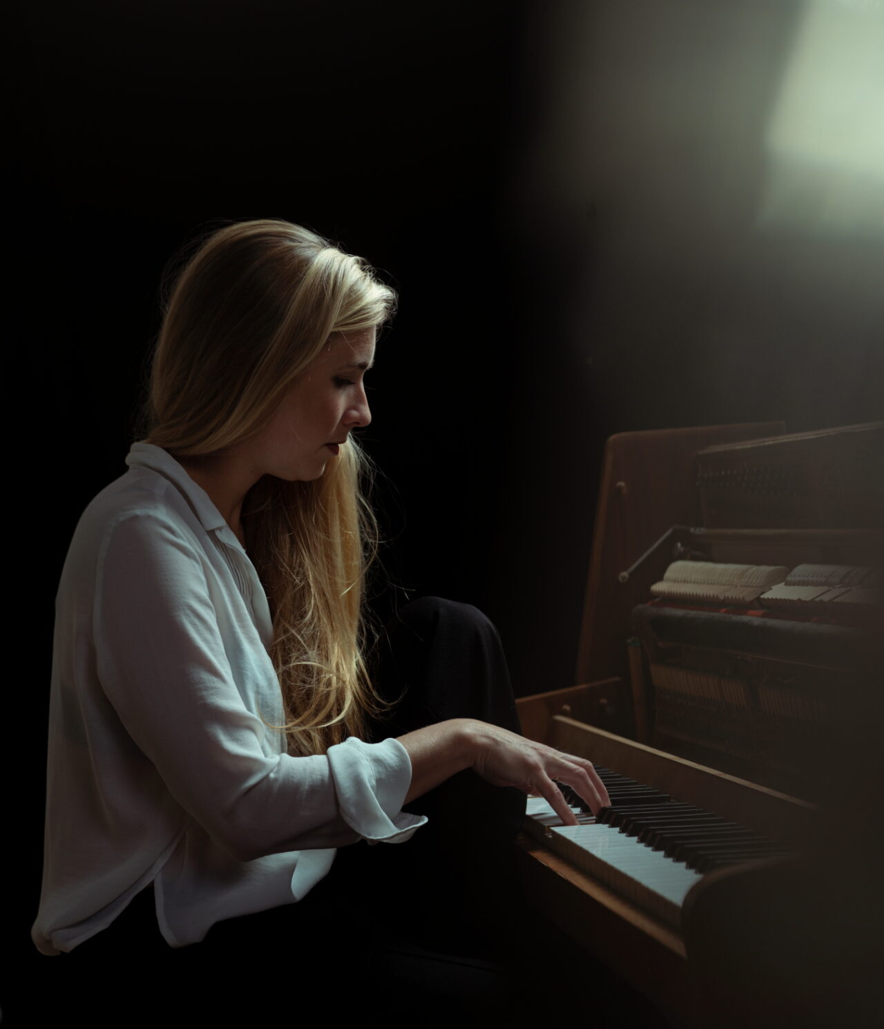 Een vrouw met lang, licht haar speelt piano in een schemerige setting met sfeervolle verlichting.