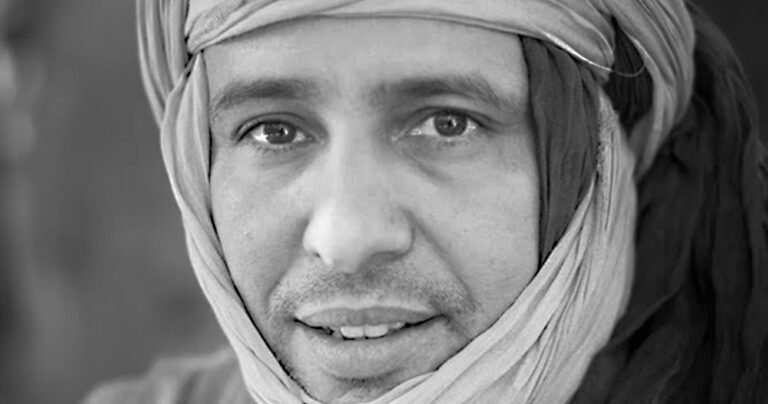 Een close-up van een man met een hoofddoek die recht in de camera kijkt.