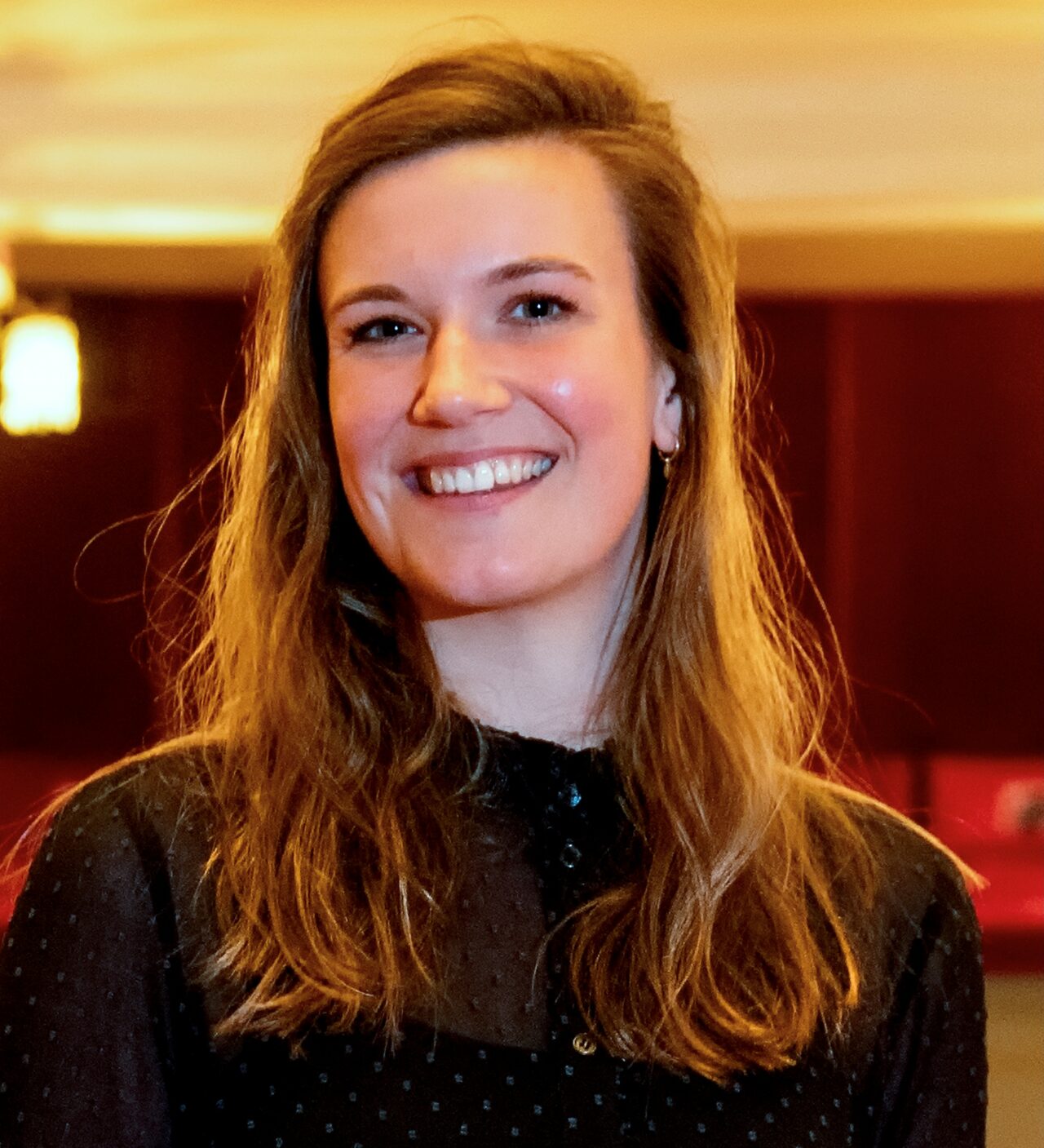 Een glimlachende jonge vrouw met lang, golvend haar, gekleed in een zwarte blouse, poseert in een stijlvolle omgeving.