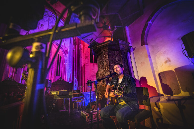 Een muzikant zit op een podium in een kleurrijk verlichte ruimte en speelt gitaar.