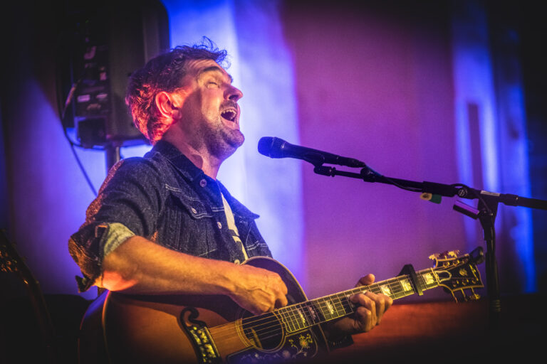 Een zanger speelt gitaar en zingt gepassioneerd op een podium met een kleurrijke achtergrond.