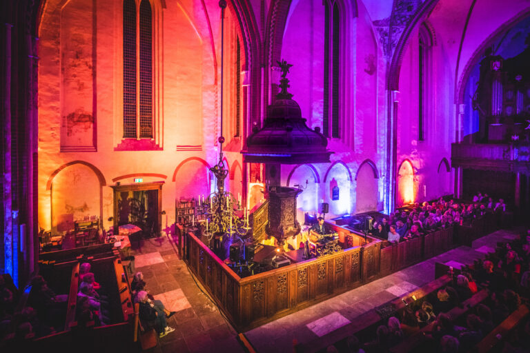 Een kerkinterieur met kleurrijke verlichting en een houten koorafscheiding, waar publiek zit te luisteren.