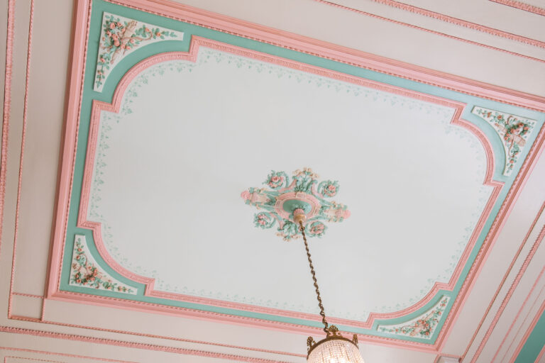 Versierde plafond met pastelgroene en roze details, inclusief een centrale ornament en een hangende lamp.