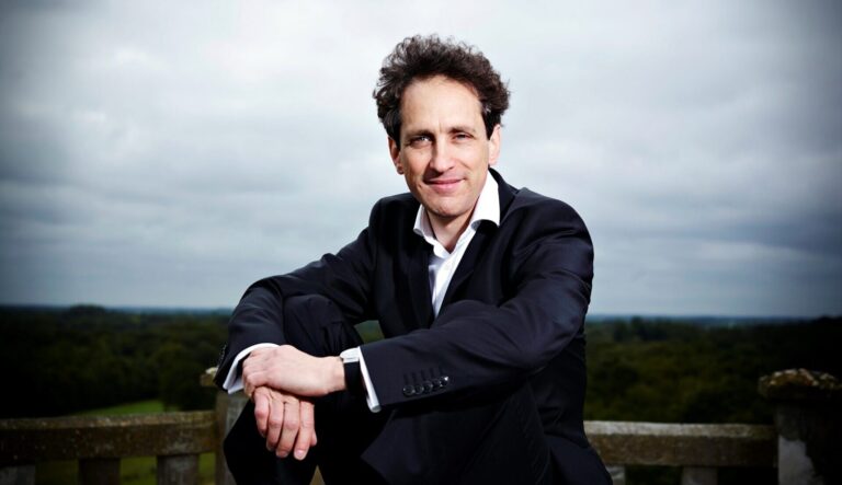 Een man in een zwart pak zit op een houten railing met een landschap op de achtergrond.