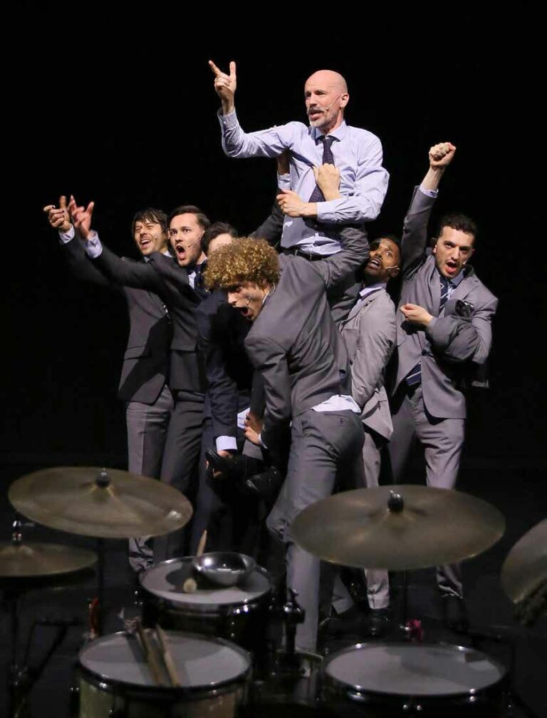 Een groep mannen in pak staat in een dynamische pose, waarbij één man op de rug van een andere man staat, met een drumstel op de achtergrond.