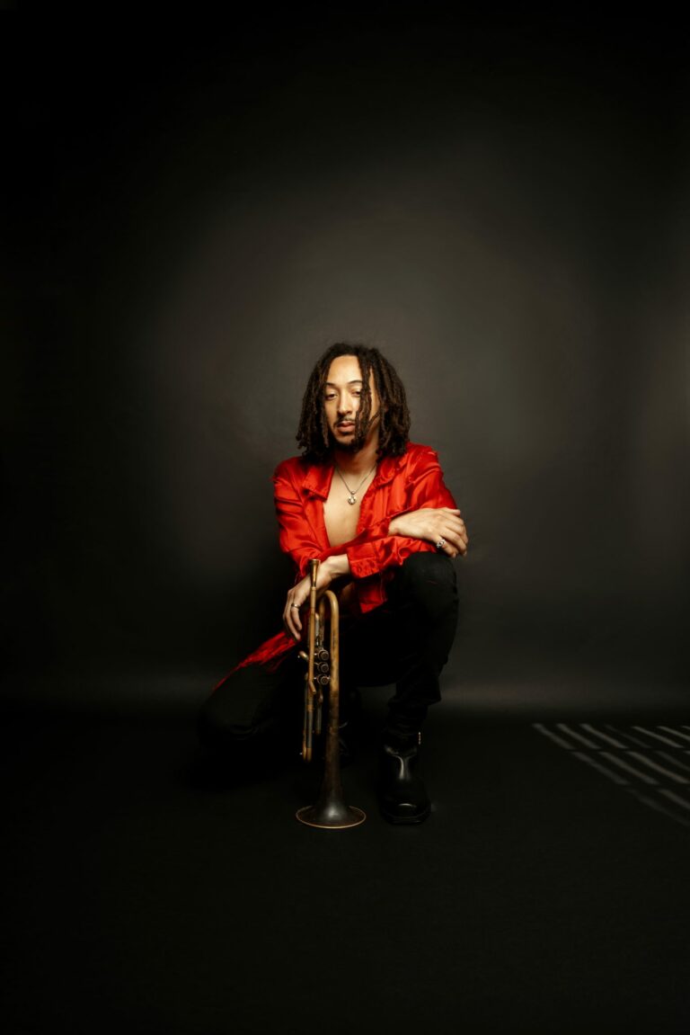 Man in a red shirt sitting on the floor, holding a trumpet in a dark setting.