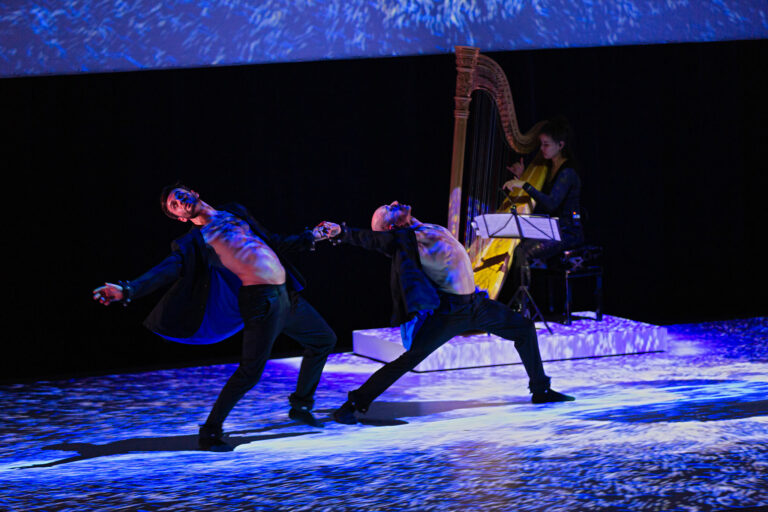 Twee dansers bewegen expressief op een podium, terwijl een harpist achter hen speelt.