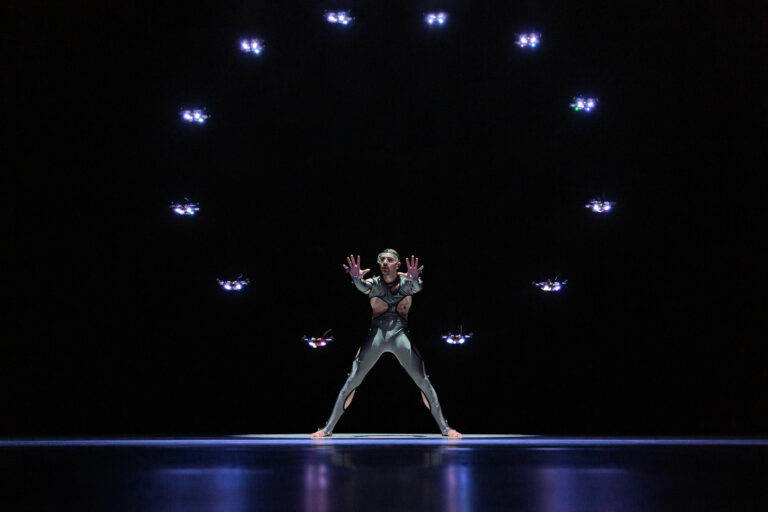 Een performer in een glanzend kostuum staat centraal op een podium, omringd door een ring van lichtpunten tegen een donkere achtergrond.