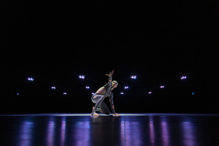 Een danser in een dramatische pose op een donkere podium met zachte verlichting.
