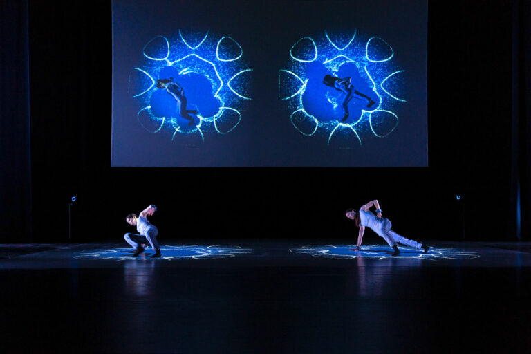 Twee dansers voeren een choreografie uit op een podium, terwijl ze worden verlicht door kleurrijke projecties op een scherm achter hen.