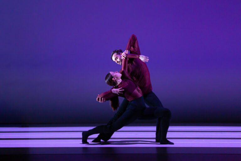 Twee dansers in rode outfits voeren een dynamische beweging uit op een gestreepte vloer met een paarse achtergrond.