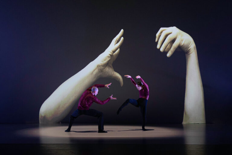 Twee dansers voeren een choreografie op voor een grote achtergrond met uitgestrekte handen.