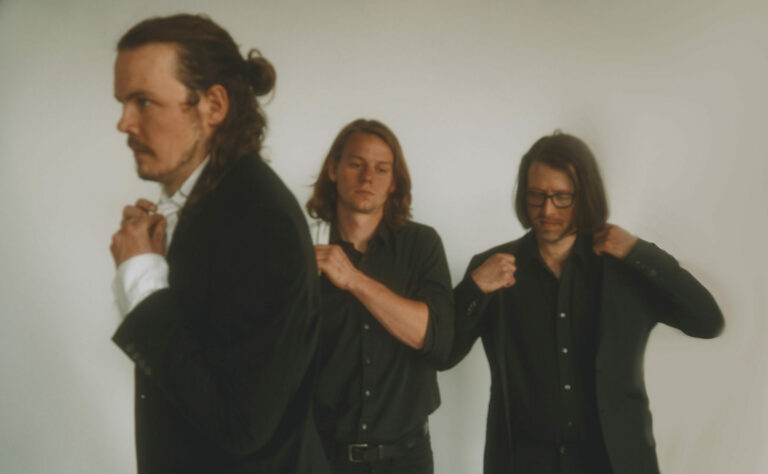 Drie mannen in zwarte outfits staan in een opstelling tegen een lichte achtergrond.