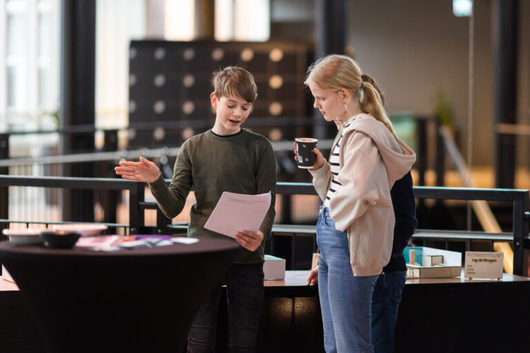 Twee kinderen staan aan een tafel in een moderne ruimte, waarbij het ene kind een document vasthoudt en het andere kind een beker koffie.