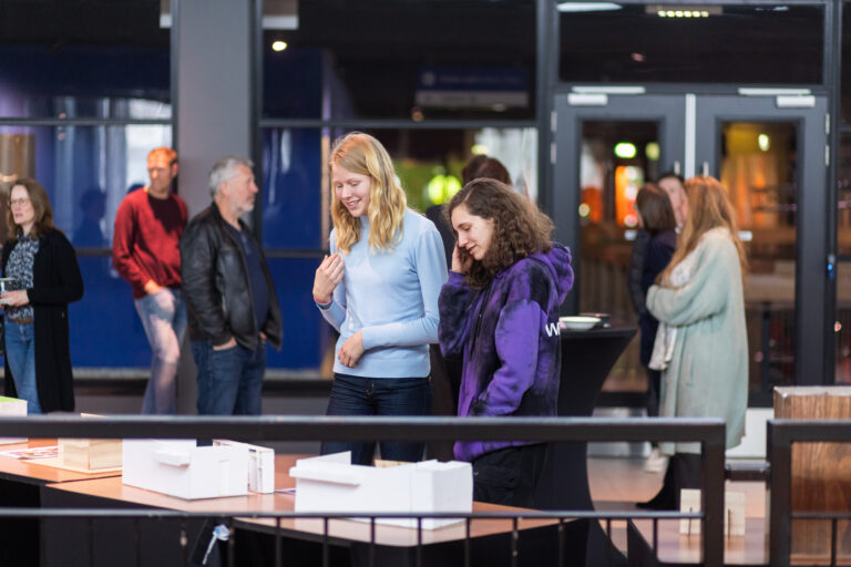 Twee jonge vrouwen bekijken modellen of kunstwerken op een tafel, terwijl andere mensen op de achtergrond staan in een drukke ruimte.