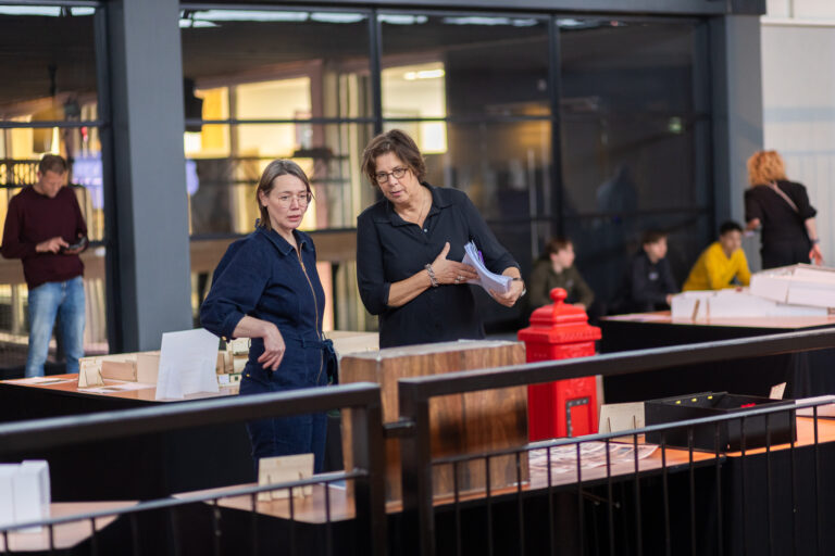 Twee mensen staan achter een houten balie in een moderne ruimte, met een rode brievenbus op de achtergrond.