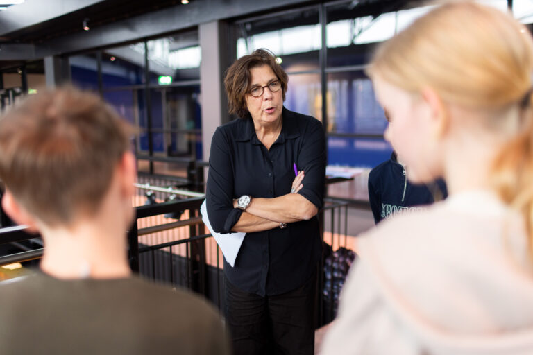 Een persoon met een korte, donkere haarsnit staat met gekruiste armen en praat met een kleine groep mensen in een expositieruimte.