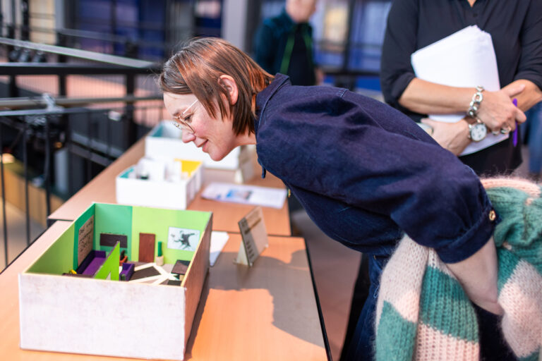 Een persoon buigt zich voorover om naar een model in een doos op een tafel te kijken, omringd door andere mensen.