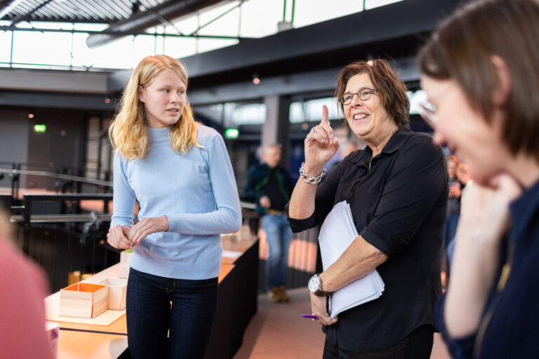 Twee mensen in gesprek in een moderne binnenruimte, waarbij de vrouw rechts zich aanspreekt en haar hand opsteekt.