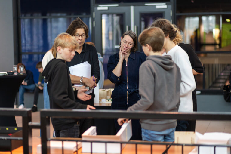 Een groep mensen, waaronder twee kinderen en drie volwassenen, bespreekt iets samen bij een tafel.