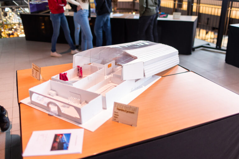 Een wit schaalmodel van een interieurontwerp ligt op een houten tafel, met bezoekers op de achtergrond.