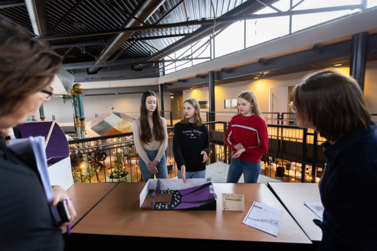 Een groep jongeren staat aan een tafel in een grote, moderne ruimte met een open houten vloer en hoge ramen.