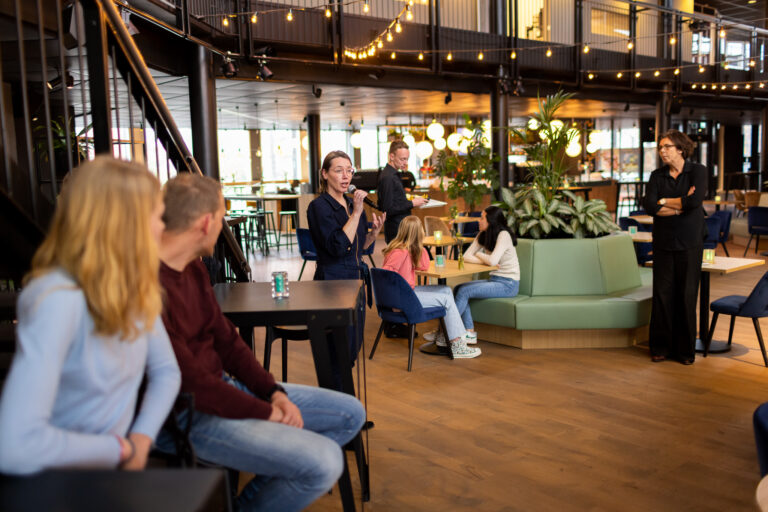 Een vrouw spreekt in een ontspannen sfeer in een café, omringd door andere mensen die aandachtig luisteren.