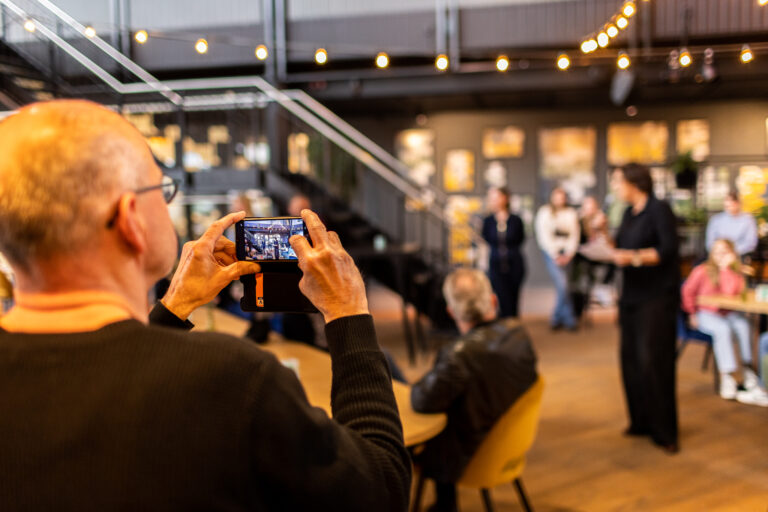 Een man maakt een foto van een evenement met een smartphone terwijl andere mensen op de achtergrond staan.