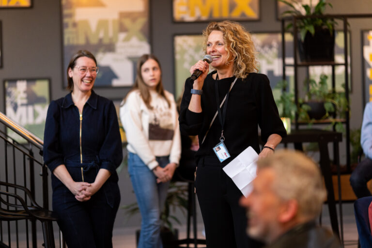 Een vrouw met een microfoon spreekt, terwijl er twee andere vrouwen naar haar luisteren in een gezellige, verlichte omgeving.