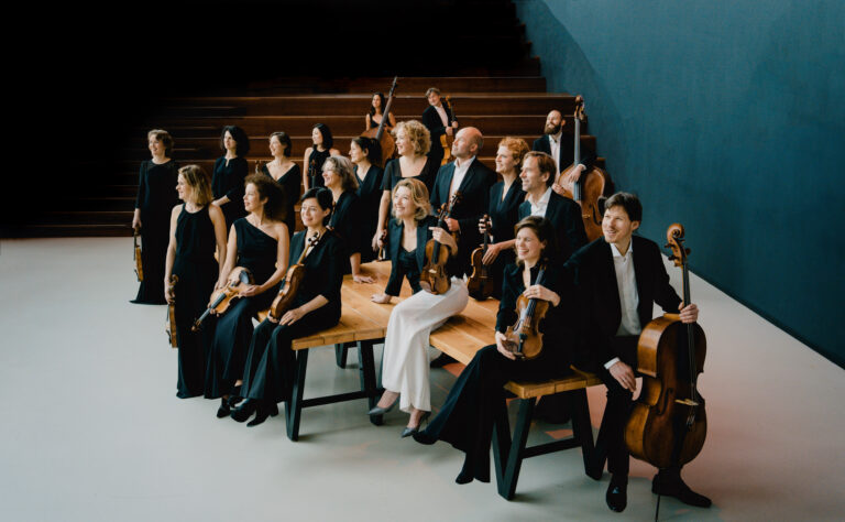 Een groep muzikanten in formele kleding staat en zit op banken in een concertzaal.