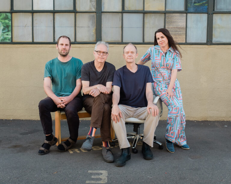 Een groep van vier mensen poseert zittend voor een gevel met grote ramen.