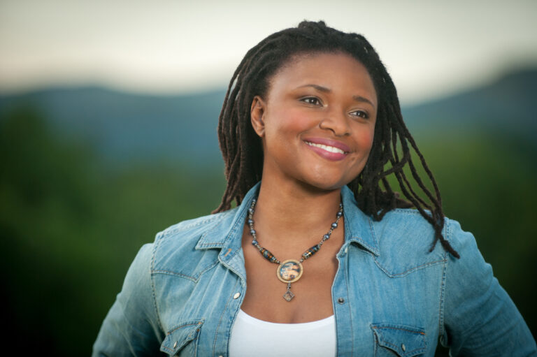 Portrait of a smiling woman wearing a denim jacket and necklace, set against a natural background.