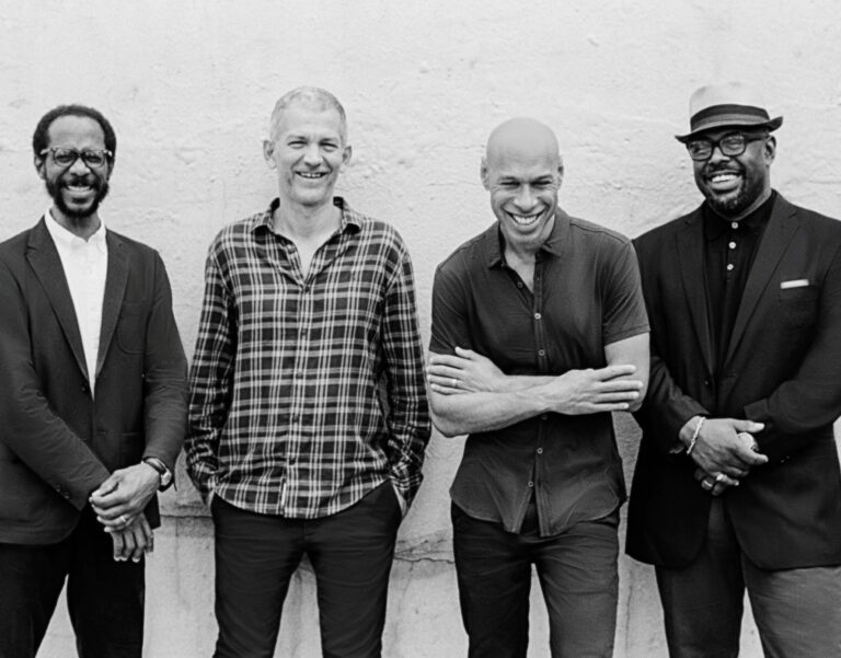 Vier mannen staan naast elkaar tegen een neutrale achtergrond, met verschillende stijlen van mode en een ontspannen houding.