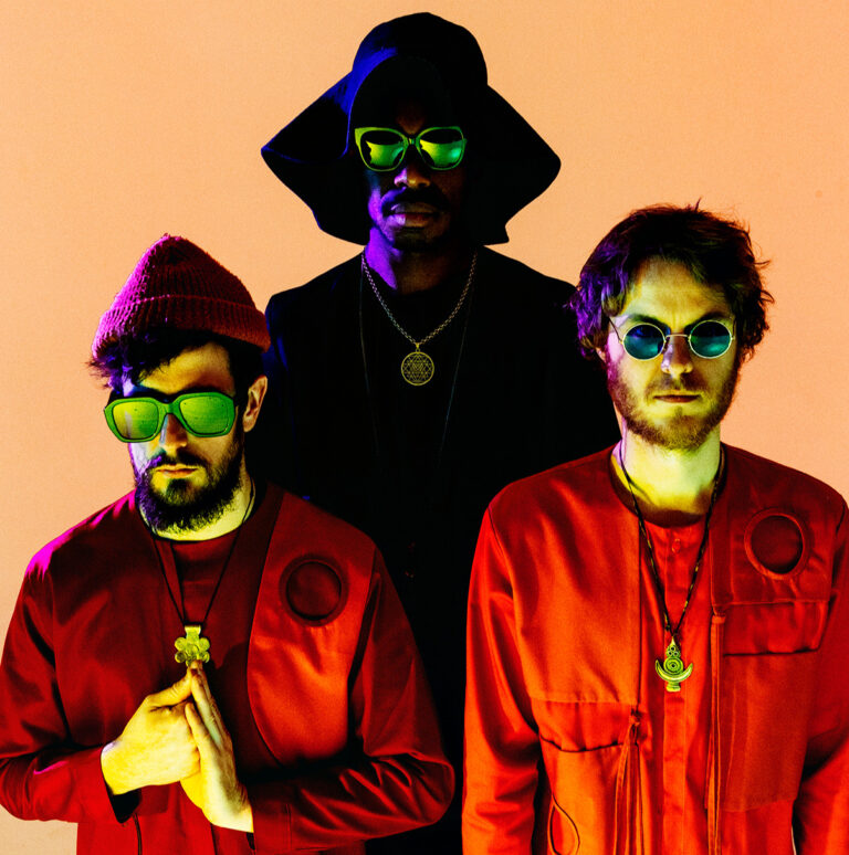 Drie mannen in kleurrijke outfits met opvallende brillen, staand voor een abstracte achtergrond.
