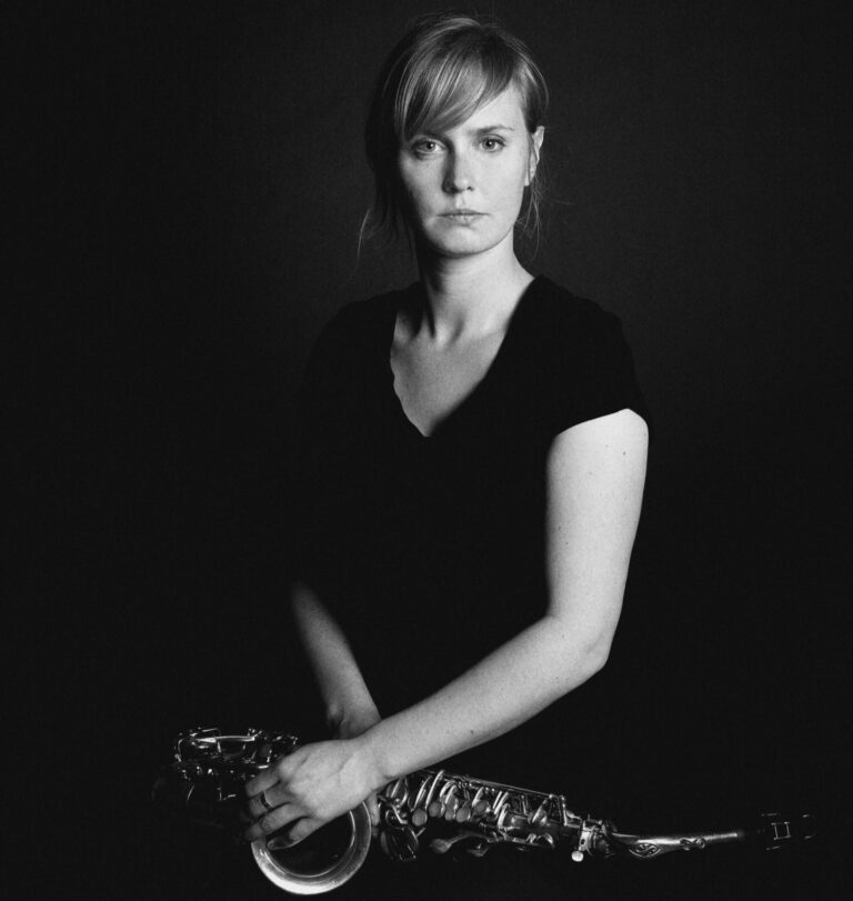 Een vrouw in een zwart shirt houdt een saxofon vast, terwijl ze recht in de camera kijkt, tegen een donkere achtergrond.