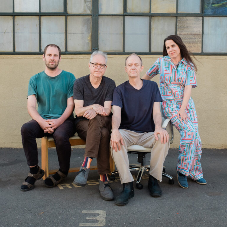 Een groep van vier mensen poserende voor een muur met grote ramen, zittend en staand in informele kleding.