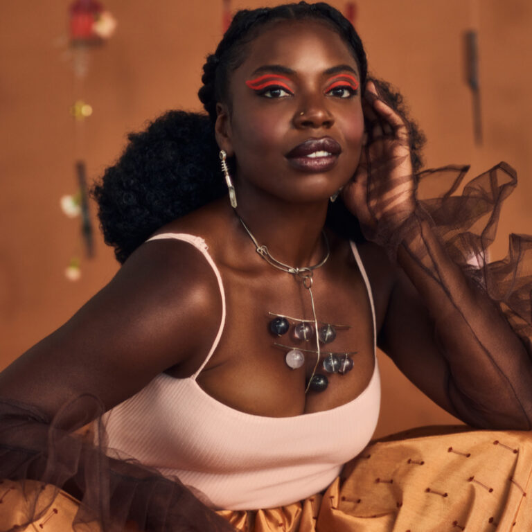 Een vrouw met een opvallende blik en kleurrijke make-up posando voor een oranje achtergrond, met een opvallende ketting en het haar in een losse stijl.