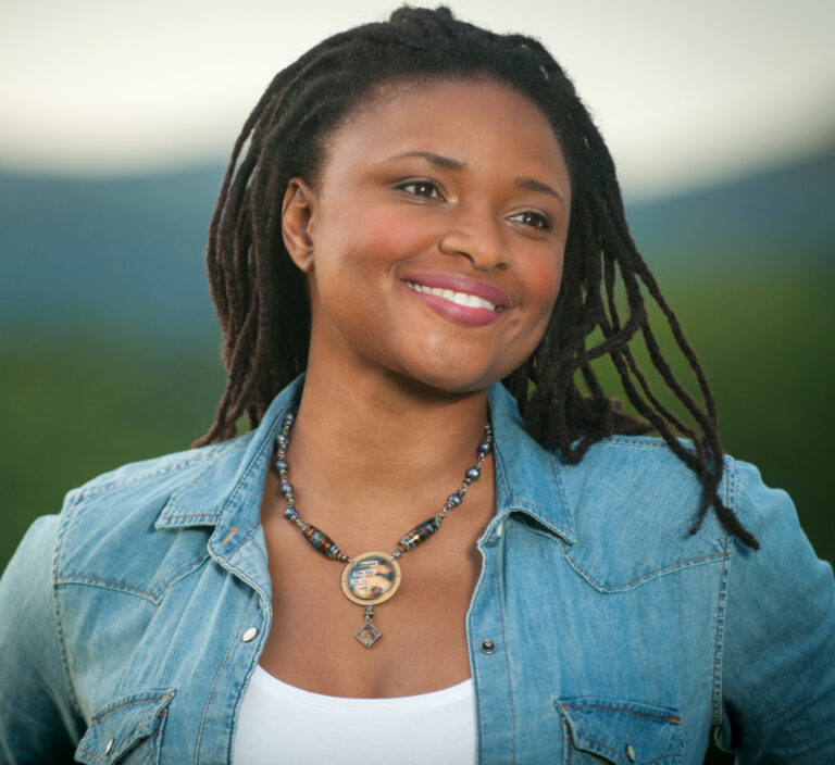 Een vrouw met een glimlach, gekleed in een spijkerjack en een ketting, poseert op een achtergrond van natuur.