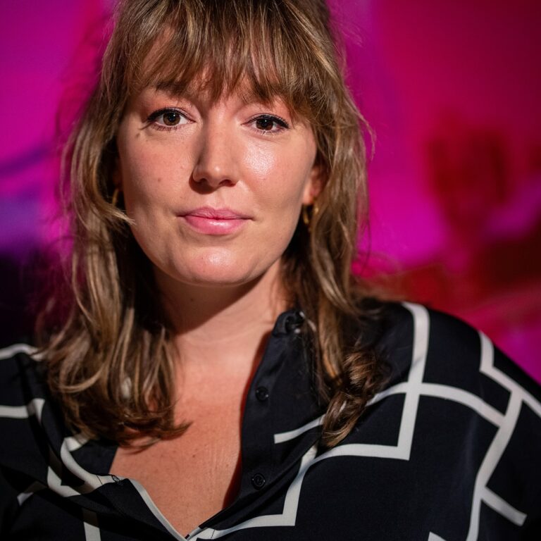 Een vrouw met bruin haar en een zwart-witte blouse poseert met een lichte glimlach tegen een paarse achtergrond.