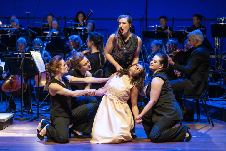 Zangeressen in zwarte kleding voeren een dramatische scène op, terwijl een zangeres in een lichte jurk centraal ligt.