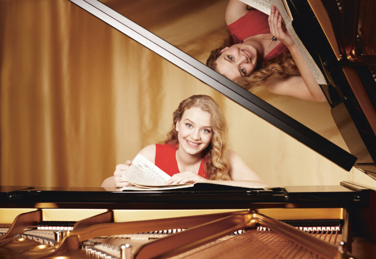 Een glimlachende vrouw in een rode jurk speelt piano, zichtbaar in de reflectie van de piano.