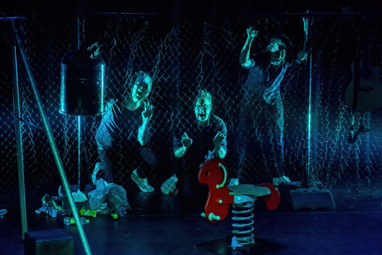 Drie mensen staan achter een gaaswand en bewegen expressief, omringd door een speelobject en kleurrijke materialen.