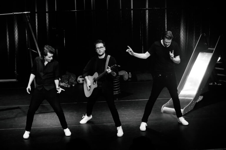 Drie muzikanten voeren een act uit op het podium, met één persoon die een gitaar bespeelt en de anderen die dansbewegingen maken.