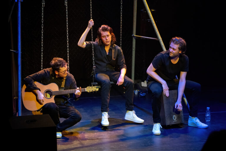 Drie muzikanten optreden op het podium, met één persoon die gitaar speelt en twee anderen die zitten en een cajón bespelen.