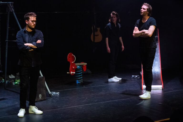 Drie performers op een donkere podium, met een gitaar en kleurrijke rekwisieten op de achtergrond.