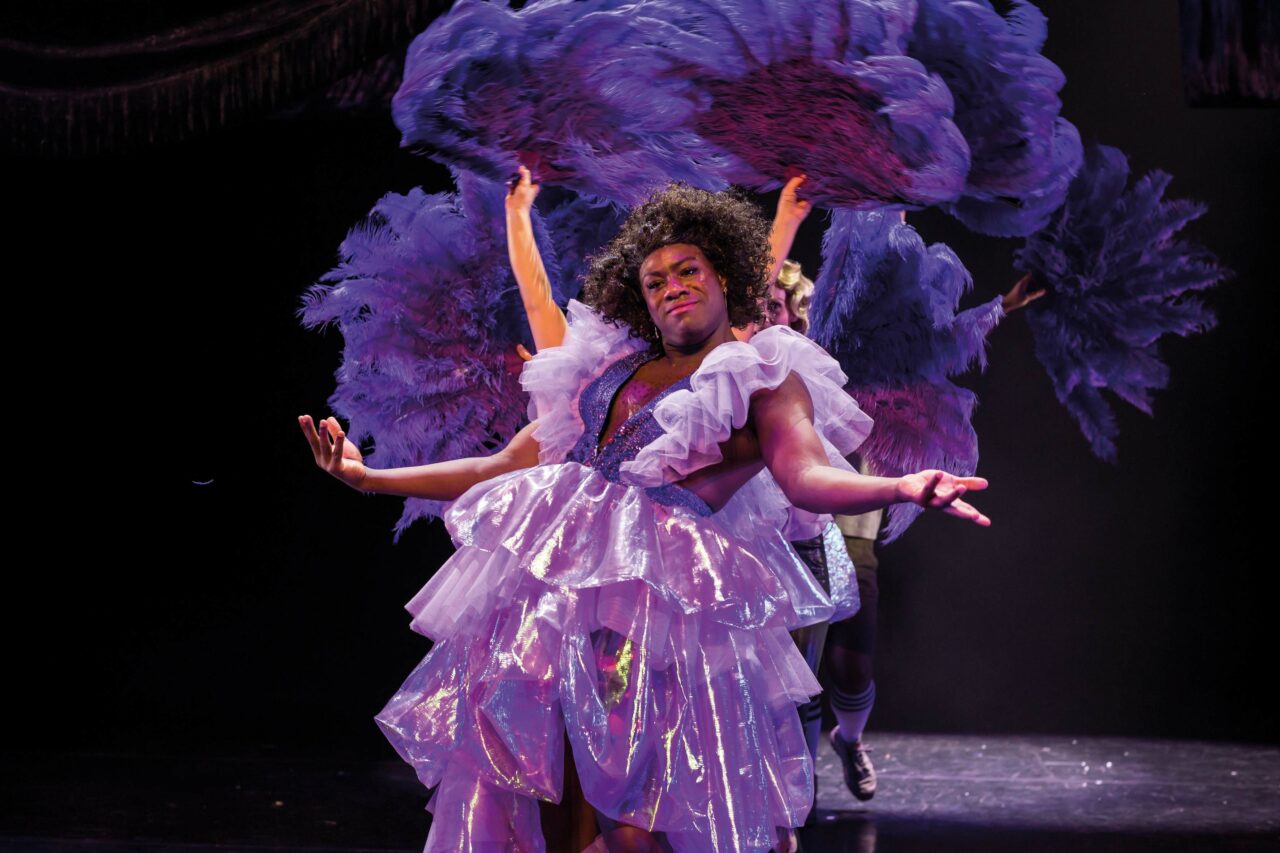 Een performer in een opvallende paarse outfit met grote pluimen, omringd door andere dansers.